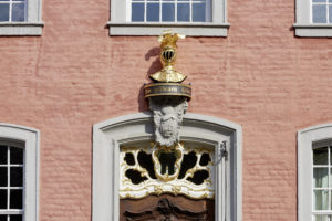 Vergoldeter Helm mit Federn über dem Eingang zum Hausteil der Familie. Darunter befindet sich der Schriftzug "Zum goldenen Helm".