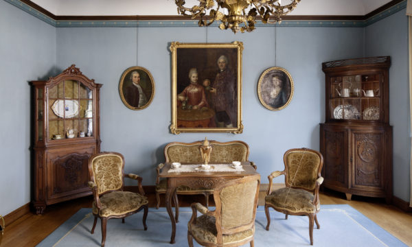 Wohnraum mit blauen Tapeten und Teppich. Auf dem Tisch der Sitzgruppe steht eine Dröppelmina, an den Wänden befinden sich Gemälde mit Familienporträts.