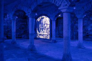 Aus Steinen und Ziegeln gemauerter Keller mit Blick durch einen Säulengang. Die Säulen tragen gemauerte Rundbögen. Der Raum ist mit blauem Licht untermalt, in der Mitte zwischen zwei Säulen ist die Projektion einer Walke auf Leinwand zu sehen.