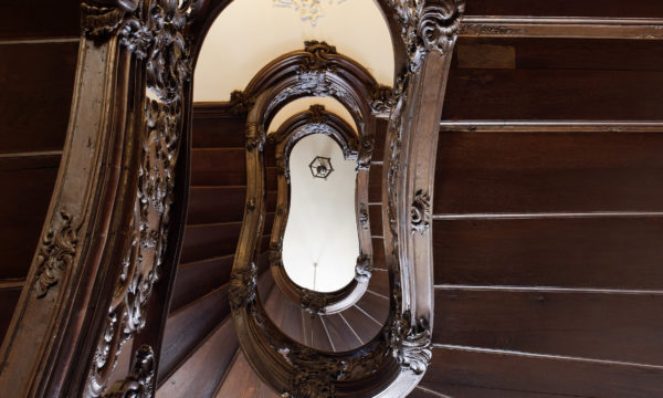 Blick von unten durch das Auge einer Treppe, deren Geländer reich mit floralen und bildhaften Schnitzereien geschmückt ist.