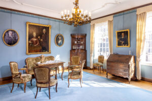Wohnraum mit blauer Wandfarbe und Teppich. Auf dem Tisch der Sitzgruppe steht eine Dröppelmina, an den Wänden befinden sich Gemälde mit Familienporträts. Aus der Mitte der Decke ragt ein 18 armiger Messingkronleuchter mit Weinlaubranken heraus. Zwei große Sprossenfenster lassen warmes Sonnenlicht ein.