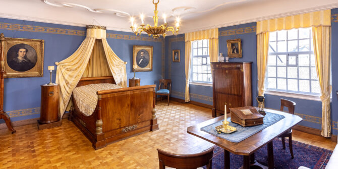 Schlafzimmer mit Himmelbett und blau gehaltenen Wänden an denen sich Porträts befinden.
