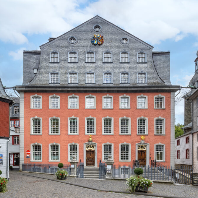Frontalansicht des Roten Hauses Monschau. Der Backsteinbau ist in den unteren drei Etagen blass rosa-rot getüncht. Zwei weitere Etagen liegen im verschieferten Mansarddach mit Kreuzgiebel. In der Dachspitze sind drei kleine runde Fenster zu erkennen, dazwischen prangt mittig das in Blau- und Goldtönen eingefärbte Doppelwappen der Scheibler und Böcking Familie. Vor dem Haus ist ein kleiner mit Eisengitter umzäunter Hof und großem Zugangstor zu erkennen. Zwei Freitreppen aus Blaustein führen zu den beiden Hauseingängen mit dunklen Eichenholztüren, über deren Oberlicht die beiden vergoldeten Hauszeichen "Zum goldenen Helm" und "Zum Pelikan" hängen. 34 große, weiße Sprossenfenster mit Blausteineinfassung sowie Ecklisenen aus Blaustein runden den edlen Eindruck ab.